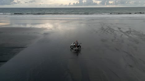 aéreo: hombre conduce una motocicleta a través de aguas poco profundas en una amplia playa de arena
