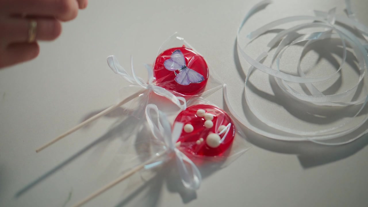 Hände schneiden vorsichtig die Verpackung um rote Lutscher. Eine Person bindet weiße Bänder um die Lutscher. Lutscher liegen auf einer weißen Oberfläche mit einem rosa Tablett in der Nähe.