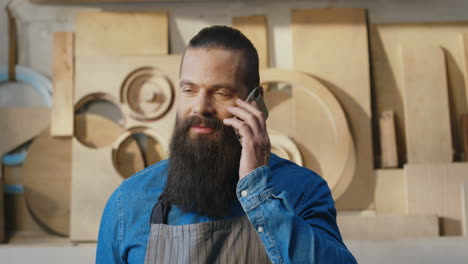 retrato de hombre barbudo caucásico en delantal hablando por teléfono y sonriendo a la cámara en el taller de carpintería