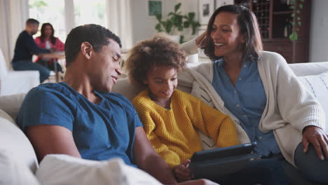 parents and pre-teen daughter sitting on sofa using tablet, grandparents at table in background