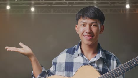 un primer plano de un adolescente asiático con una guitarra sonriendo y señalando hacia el lado en el escenario