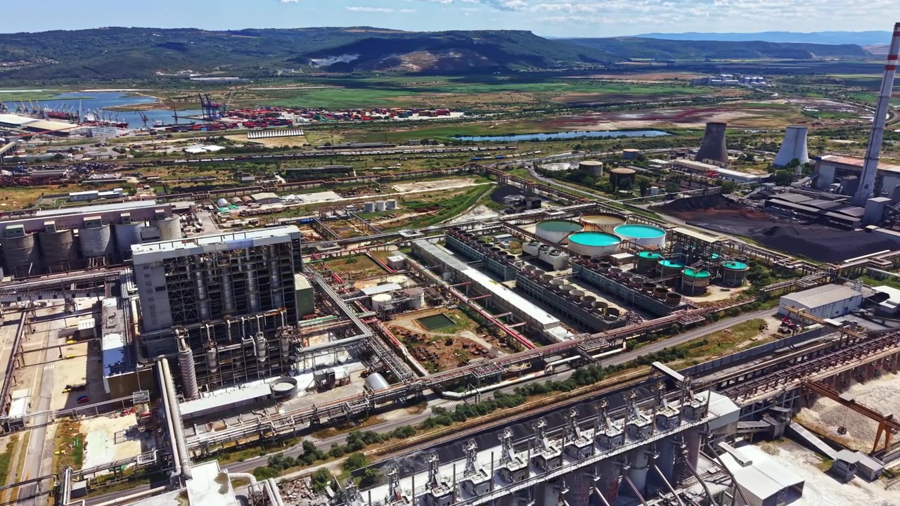 Sitio industrial en una ciudad con fábricas, tanques de almacenamiento y terreno abierto cerca de un río y montañas durante el día.