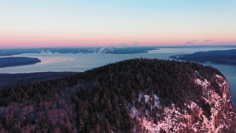 Vista-Aérea-Volando-Lejos-De-La-Cima-De-Una-Montaña-Frente-A-Un-Acantilado-Mientras-Ve-El-Lago-Helado-Y-Brumoso-Detrás-De-él-Durante-Un-Amanecer-De-Invierno