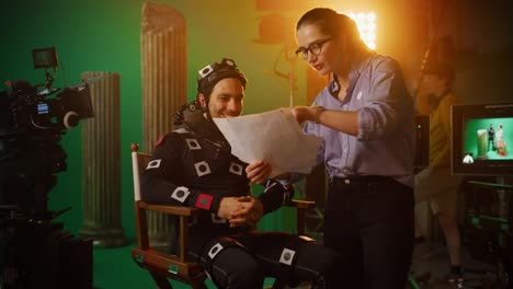 on film set: prominent female director explains scene to male actor wearing motion capture suit and playing green screen scene in superhero movie. on big film studio professional crew