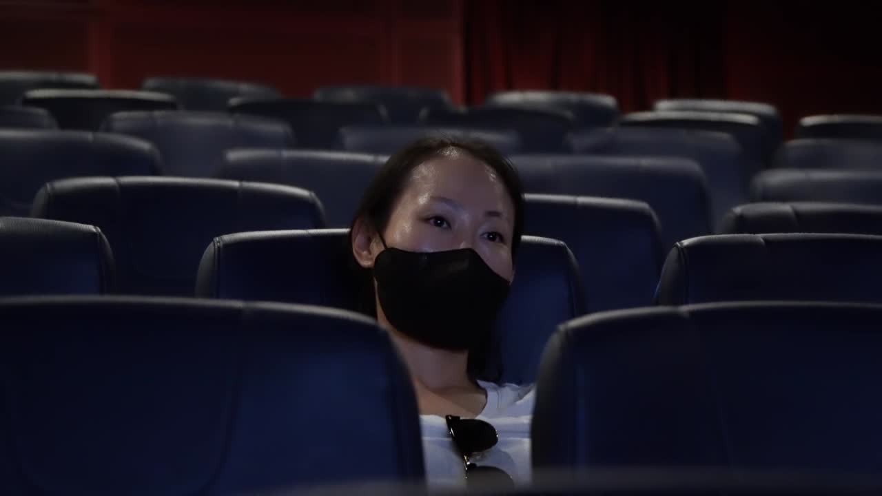 Asian Woman In Mask Sitting In An Empty Cinema And Watching Movie Alone. medium shot