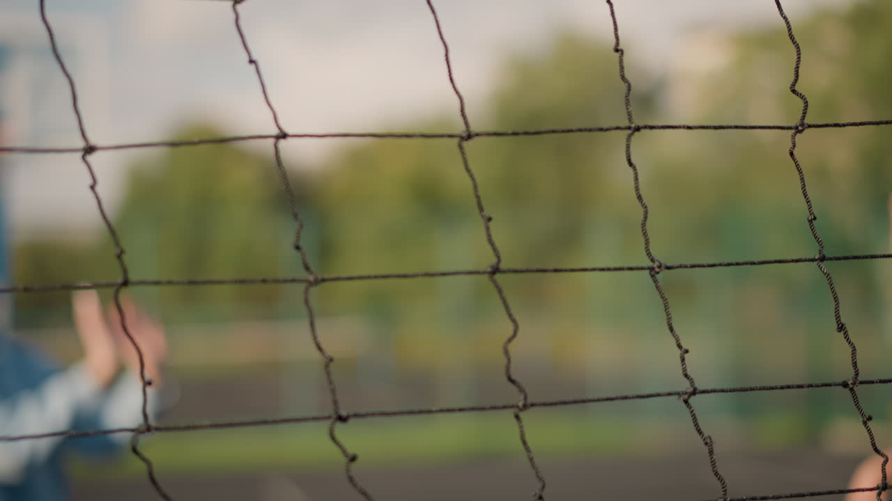 Close-up of volleyball net with blurred background of two people playing volleyball, sunlight creates golden glow on greenery, enhancing energetic outdoor scene of sports activity