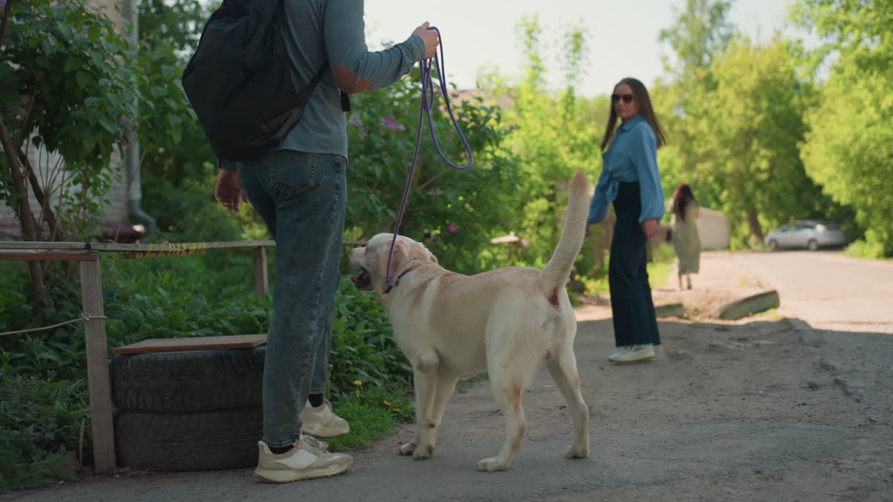 Pareja blanca con labrador se acerca a un antiguo pozo de piedra en un sendero del pueblo; el perro olisquea el agua y explora; mochilas y ropa casual; entorno rústico y verde, y tranquila curiosidad durante una excursión por el barrio