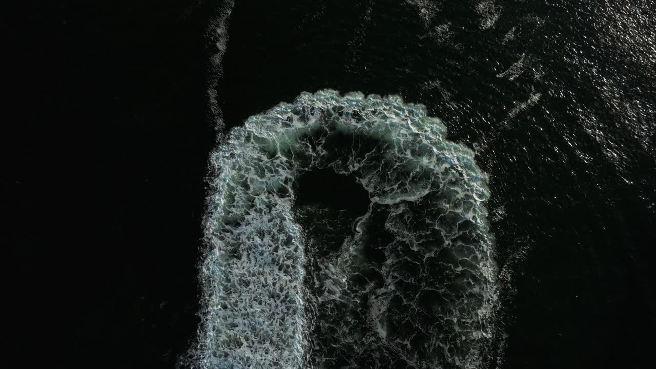 A red boat swiftly maneuvers through water, leaving behind a spiral pattern of white foam and waves under a clear blue sky. The scene captures the thrill of boating