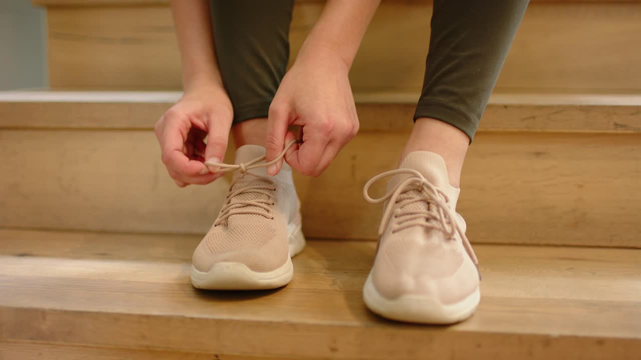 Donna africana seduta su delle scale di legno chiaro, che si allaccia i lacci allentati della scarpa da ginnastica sinistra, preparandosi a muoversi.