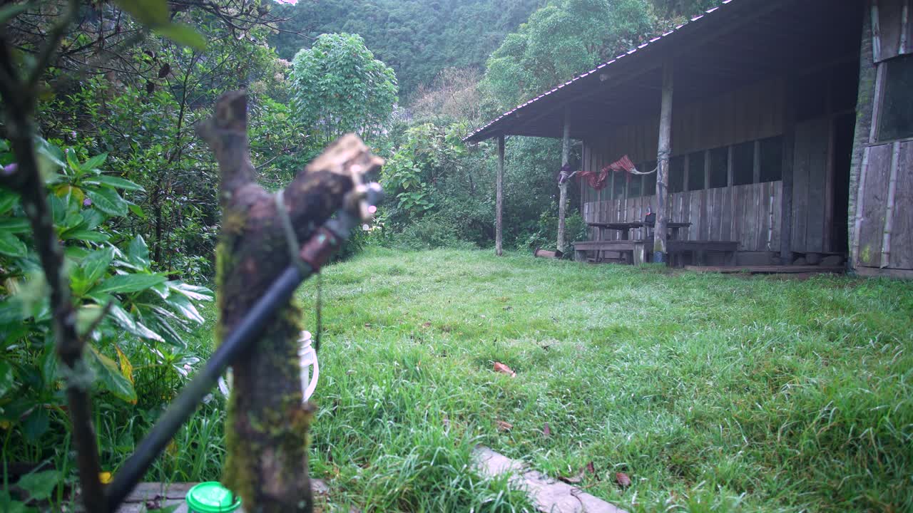 Static shot of a rustic wooden cabin surrounded by lush vegetation, with a blurred water tap in the foreground