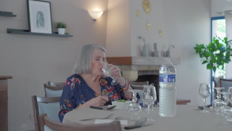 senior woman sitting alone at empty dining table, drinking water out of glass, modern indoor retirement home