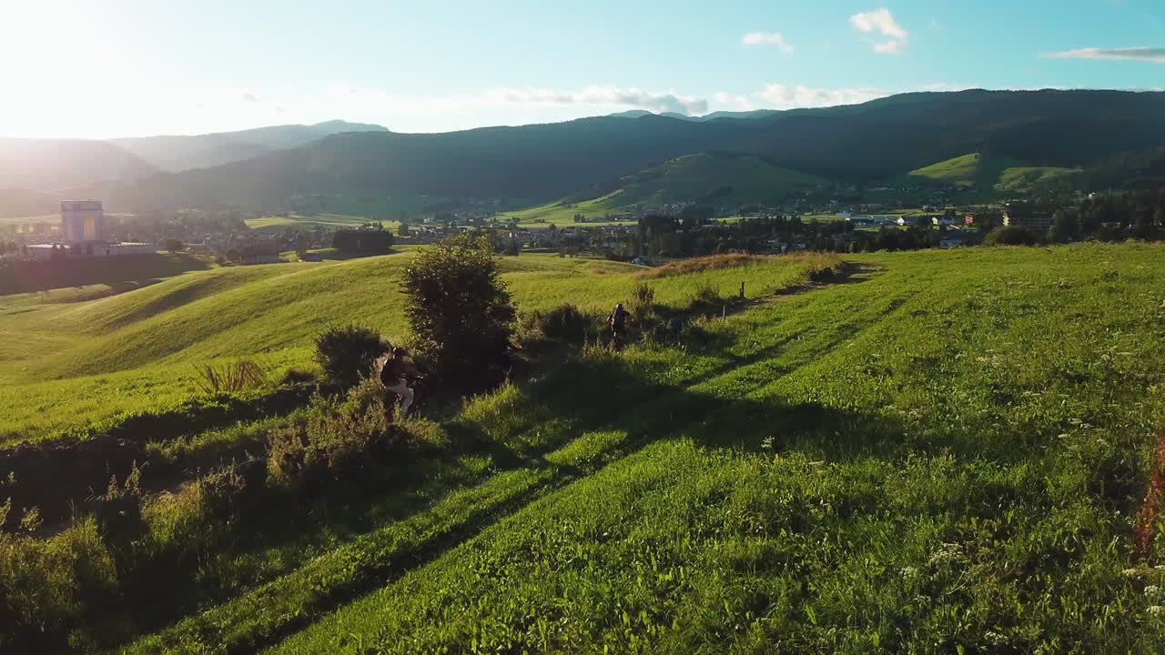 Aerial side view of mountain bikers on a dirt trail in the mountains. People on bicycles riding through grassy hills with a view of beautiful valley