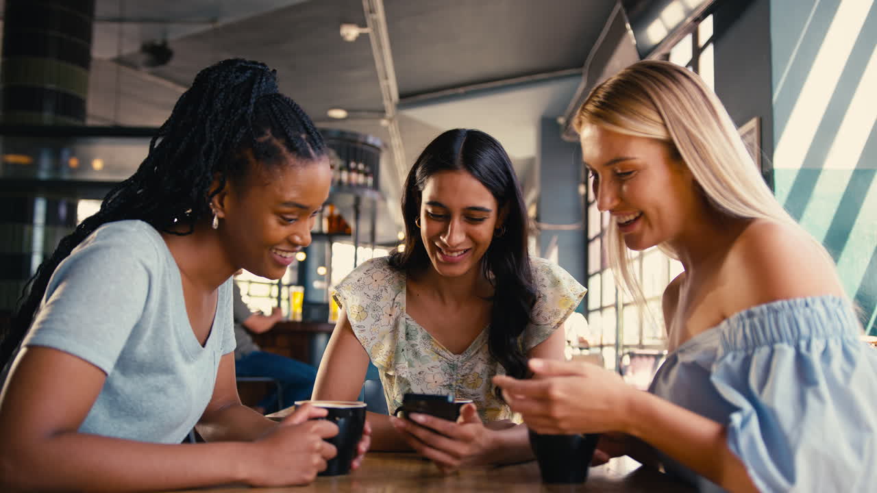 Group Of Female Friends Meeting Up In Restaurant Or Coffee Shop Looking At Mobile Phone
