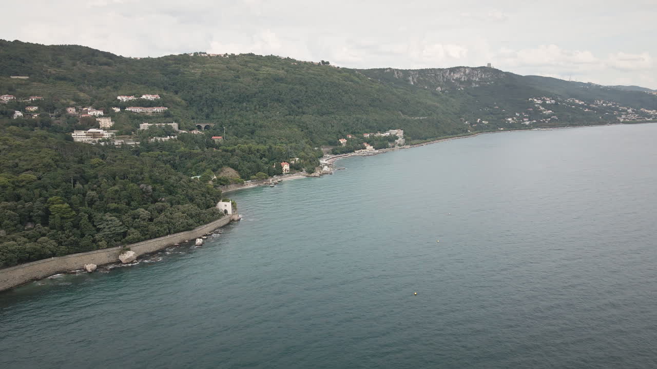 Un drone 4K UHD scorre fluidamente in avanti lungo una costa rocciosa con spiaggia, strada costiera e colline verdi sotto nuvole soffici, perfetto per film di viaggio, spot pubblicitari sul turismo, introduzioni