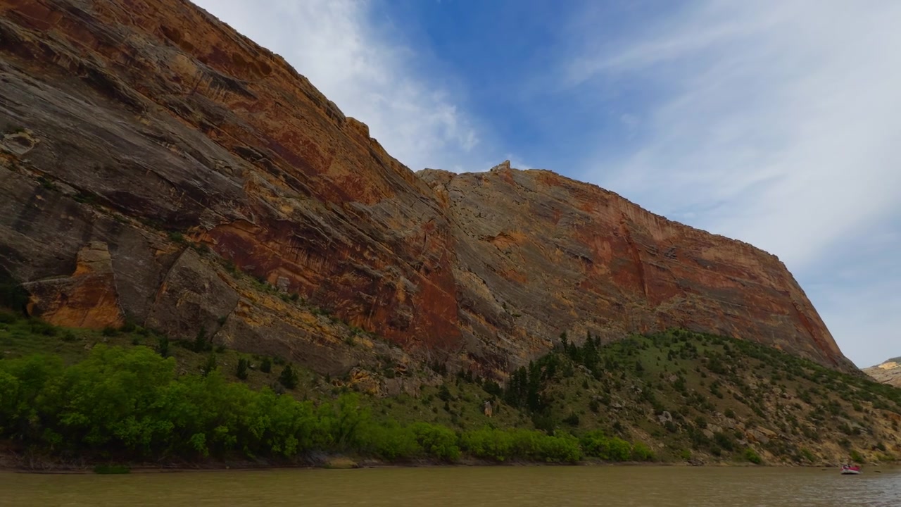 Collina di canyon stratificata e colorata si erge sopra la densa vegetazione ripariale e il fiume, panoramica a destra.