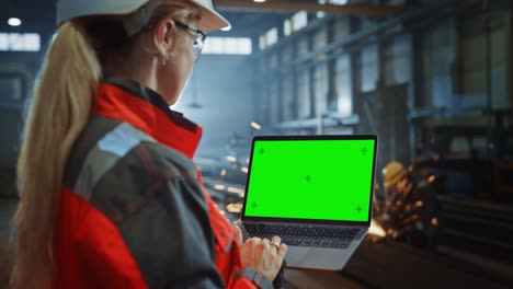 ingeniero profesional de la industria pesada utiliza una computadora portátil con pantalla verde. especialista industrial femenina trabajando en un almacén de fabricación de metal con chispas en el fondo.