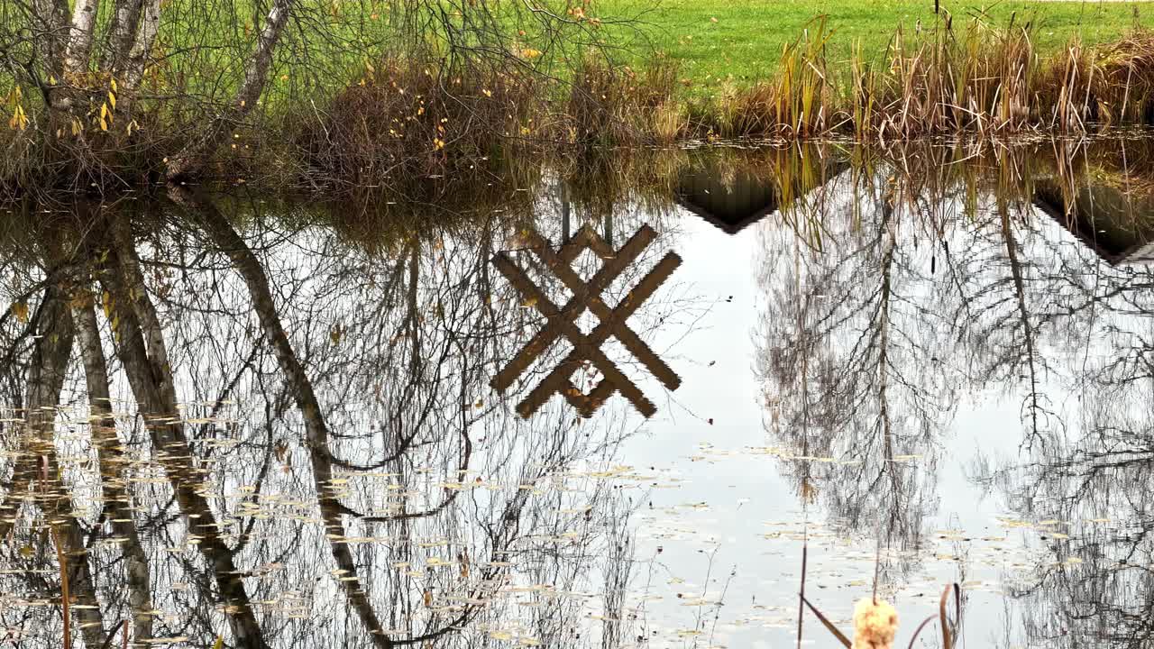 Reflective surface of pond with Latvian folk symbol artwork and leafless trees in still water