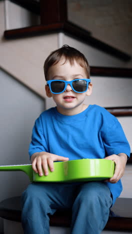 adorable caucasian baby boy sitting on the stairs holding a guitar. portrait of a lovely kid wearing sunglasses. vertical video.