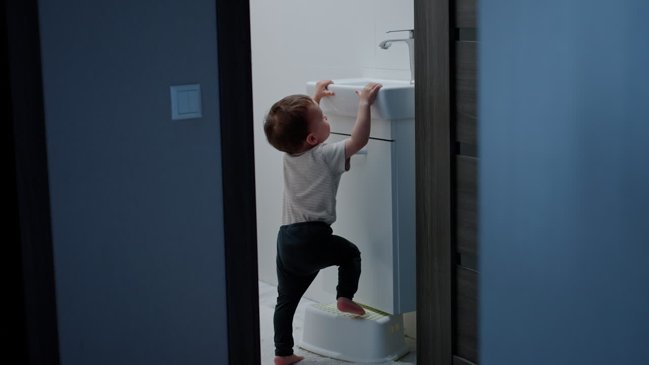 Cute baby boy takes a stand out of the chest. Smart kid stands on the prop to reach the tap.
