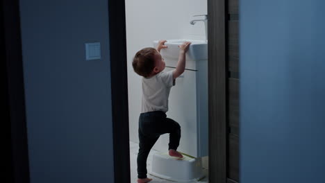 cute baby boy takes a stand out of the chest. smart kid stands on the prop to reach the tap.