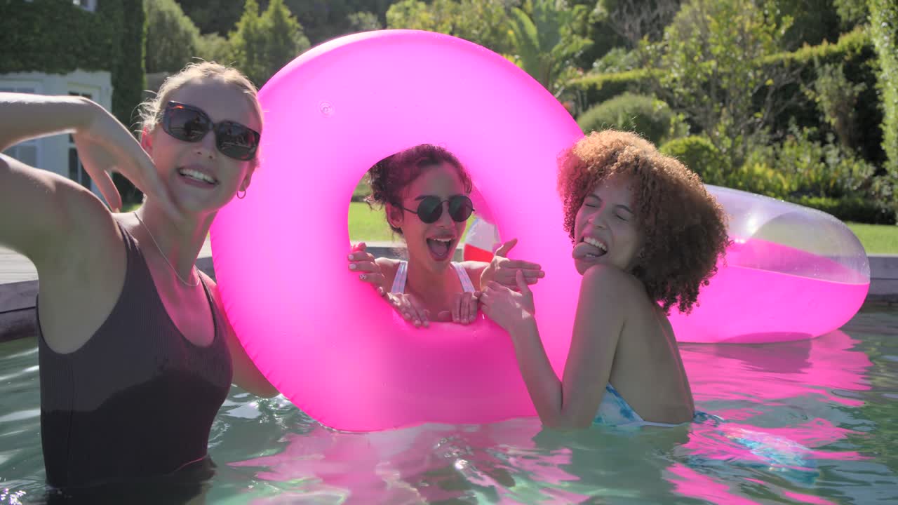 3 female friends posing for camera in swimsuits holding hands across pink donut at backyard pool