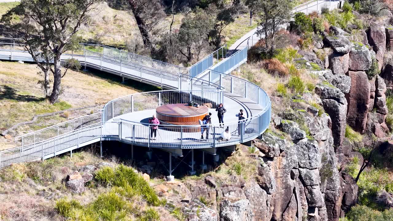 A group of people walk and pause on a modern clifftop lookout platform, surrounded by rocky terrain and bushland, under bright natural daylight. The camera remains steady, capturing an elevated wide view