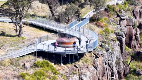 a group of people walk and pause on a modern clifftop lookout platform, surrounded by rocky terrain and bushland, under bright natural daylight. the camera remains steady, capturing an elevated wide view