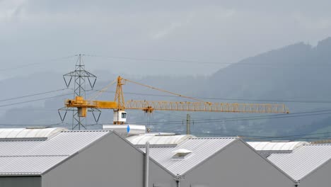 yellow construction crane with electricity pylon in background at warehouse industrial site on rainy overcast day