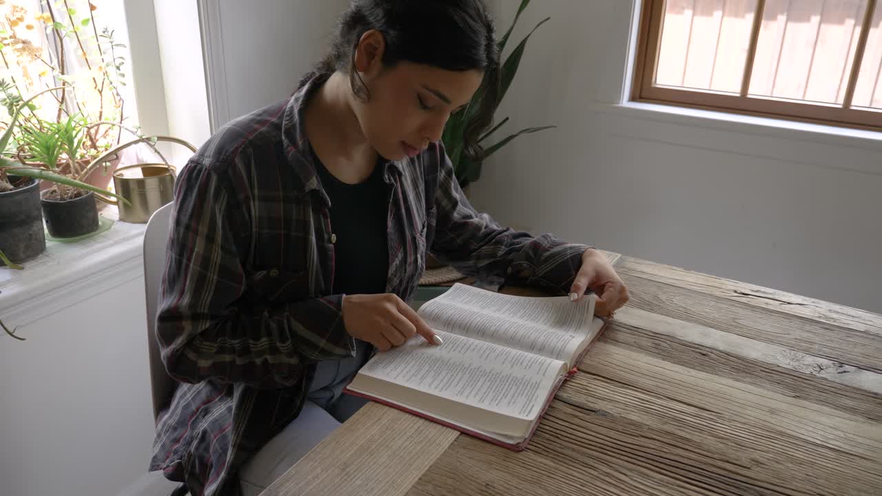 cómoda chica puertorriqueña leyendo la biblia relajándose en una mesa de madera con una ventana brillante