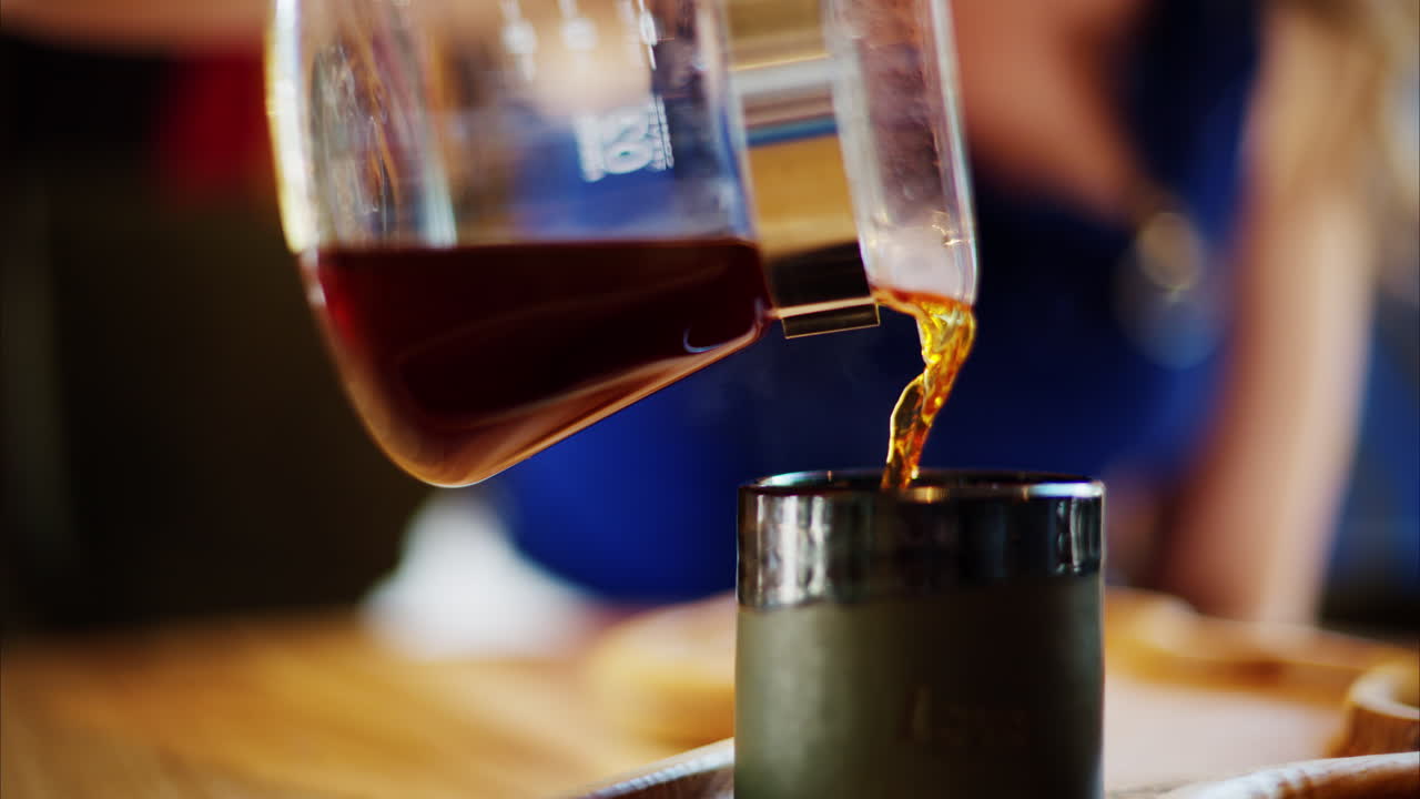 Close up of a woman pouring coffee in a cup from a glass pot standing on a wooden tray