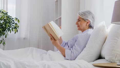 Mujer-Mayor-Leyendo-Un-Libro-En-La-Cama-En-El-Dormitorio-De-Su-Casa.-Tecnología,-Concepto-De-Vejez-Y-Personas.-Mujer-Mayor-Leyendo-Un-Libro-En-La-Cama-En-El-Dormitorio-De-Su-Casa.