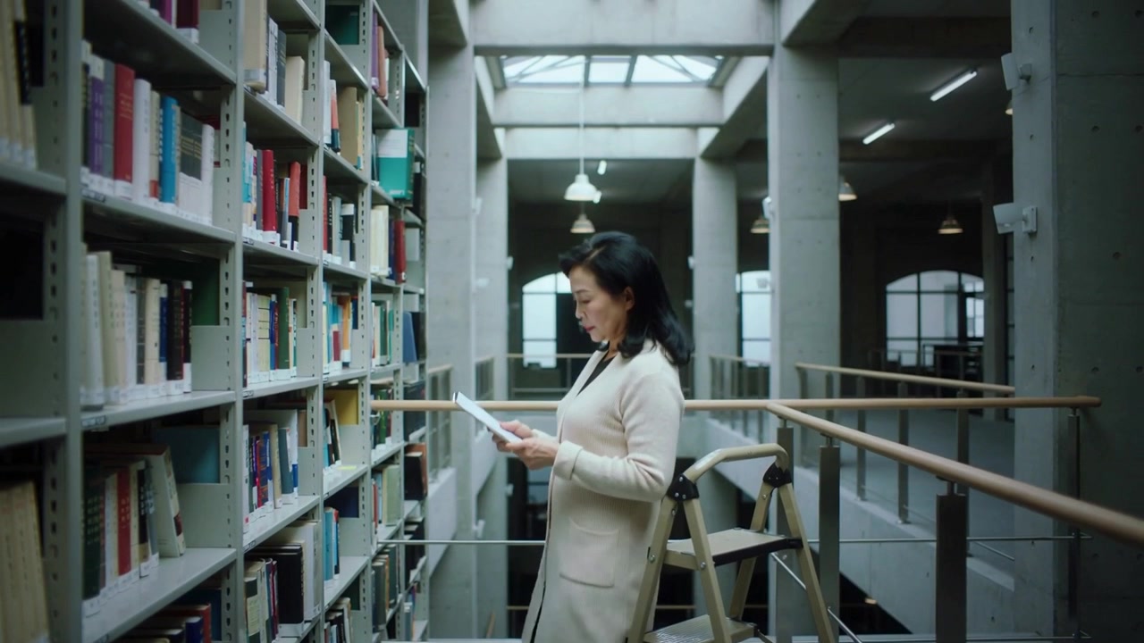 Donna che cerca, appoggiata a una scala sul soppalco di una biblioteca, che prende un libro e ne legge le pagine, indossando un cardigan.