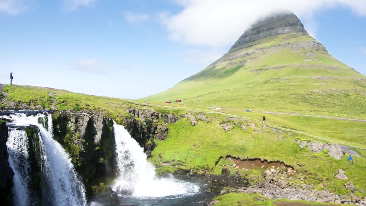 kirkjufell y cascada kirkjufellfoss en islandia, con gente caminando sobre la cascada en un cielo azul día soleado y una nube sobre la montaña