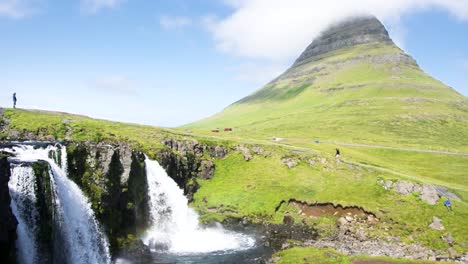 kirkjufell, and kirkjufellfoss waterfall in iceland, with people walking atop the waterfall on a blue sky sunny day and a cloud over the mountain