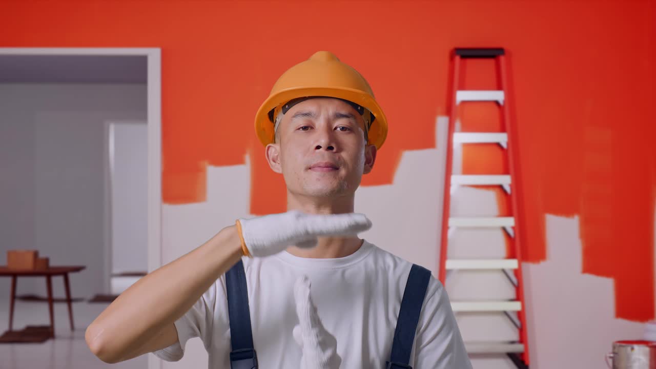 Close Up Of Asian Man Painter Wearing Safety Helmet Looking At Camera And Showing Time Out Hands Gesture While Standing With Painting Wall In Room