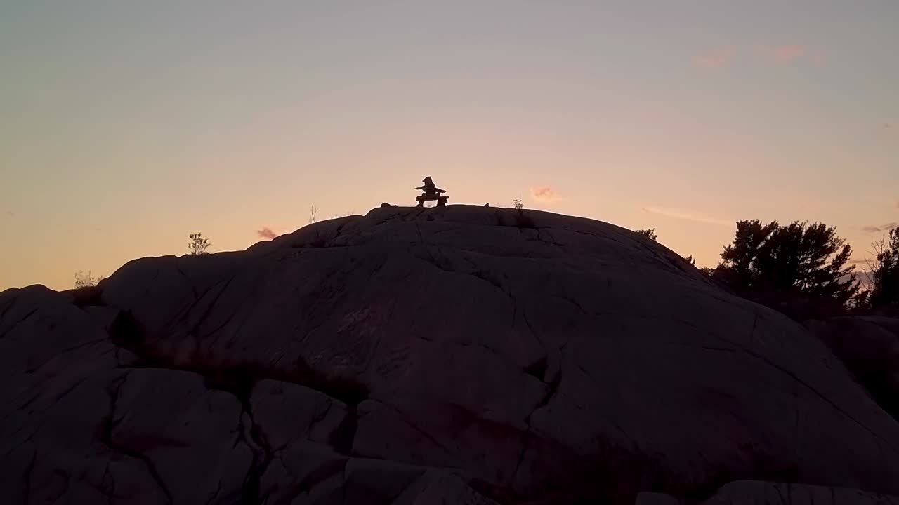 Sorvolo di Inukshuk sull'isola rocciosa di pini al tramonto, ripresa aerea con drone, dolly in ampio