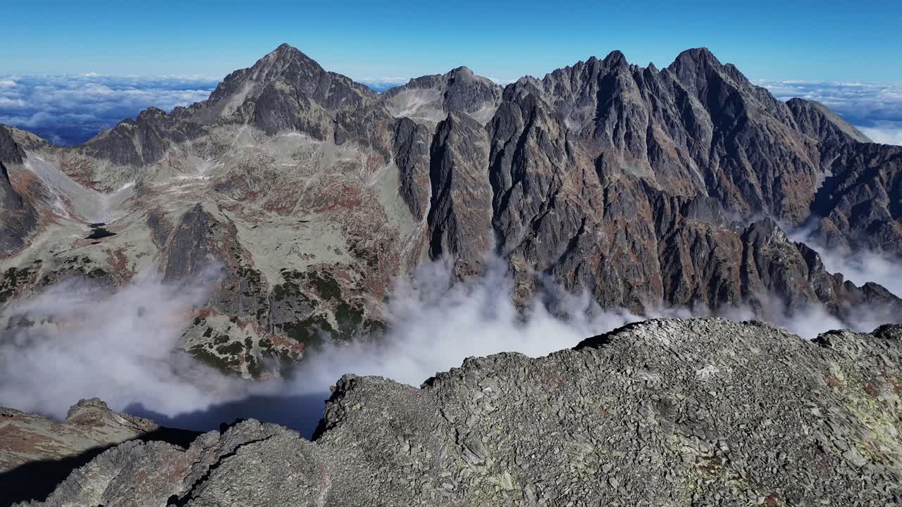 Side-flying drone shot capturing the rugged ridge of Slavkovský štít in the High Tatras, with dramatic peaks and a dense clouds below under clear blue skies