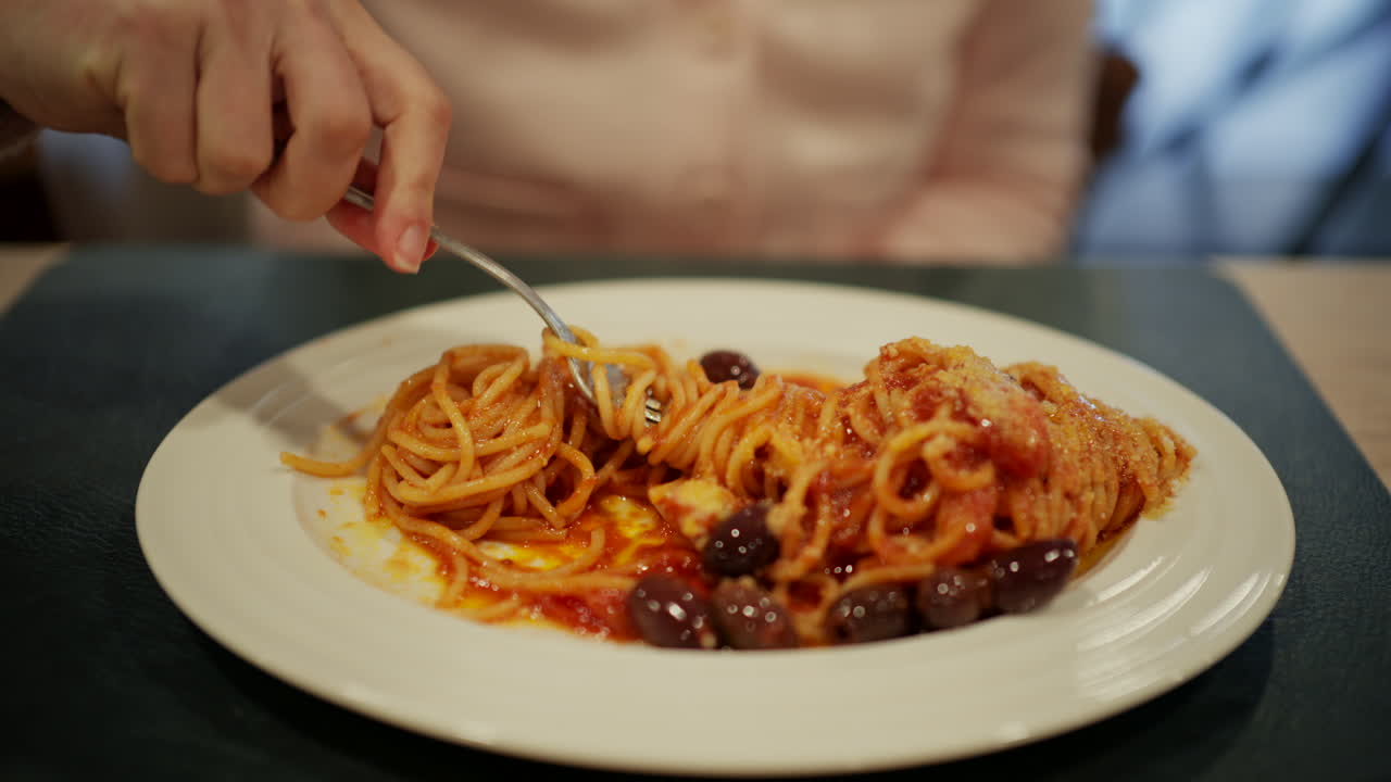 Primo piano di una persona che avvolge spaghetti alla puttanesca con una forchetta, mostrando un ricco sugo di pomodoro, olive e formaggio grattugiato