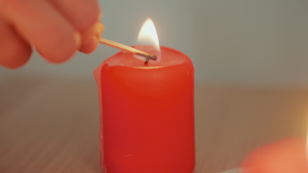 Mano che accende un fiammifero a una candela, sequenza ravvicinata dell'accensione del fiammifero e della fiamma che prende lo stoppino di una candela rossa a colonna su un tavolo di legno, movimento attento e deliberato suggerisce un rituale domestico, un allestimento per aromaterapia o un preludio.