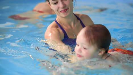 Happy-middle-aged-mother-swimming-with-cute-adorable-baby-in-swimming-pool.-Smiling-mom-and-little-child,-newborn-girl-having-fun-together.-Active-family-spending-leisure-and-time-in-spa-hotel.