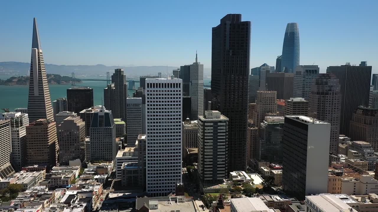 Dynamic aerial footage showcases San Francisco's downtown harbor under overcast skies, highlighting ships docked beside industrial buildings, a lush hill backdrop, and urban skyline