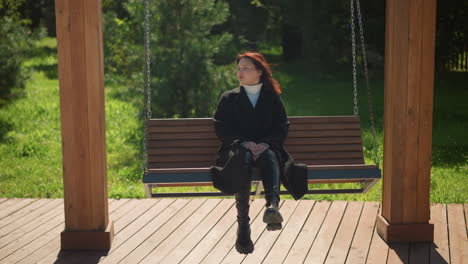 young woman in black coat sits serenely on wooden swing in sunlit park, hands between legs, gazing into the distance, surrounded by greenery