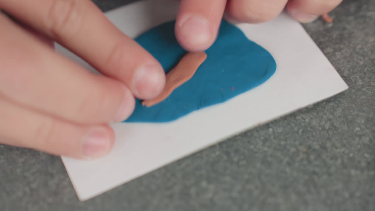 Child hands pressing clay on blue mat, shaping brown strip into pencil form, closeup focus on fingers, fine motor practice, classroom craft preparing for back to school, calm focused atmosphere