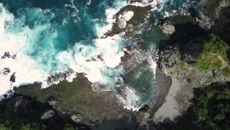 sliding overhead shot of waves of indian ocean hitting boulder and rocks in the beach in sunny condition weather with hill overgrown by trees - pengilon hill, indonesia, asia