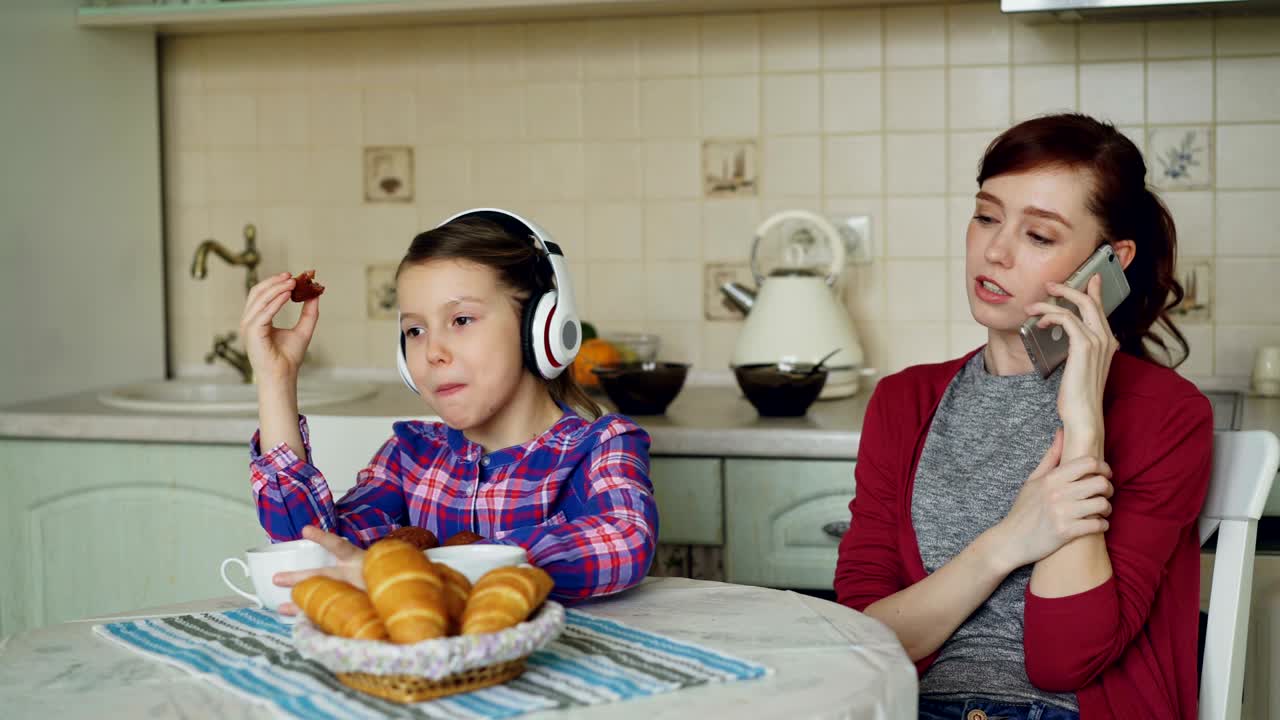 madre joven y hija linda tomando el desayuno por la mañana en la cocina en casa. niña escuchando música con auriculares comiendo muffins mientras su madre ocupada hablando por teléfono