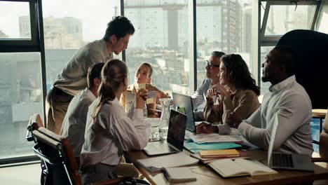 A-happy-group-of-office-workers-sit-at-a-table-and-share-among-themselves,-on-the-table-laptops-and-papers-in-an-office-with-large-windows-and-a-view-of-the-city