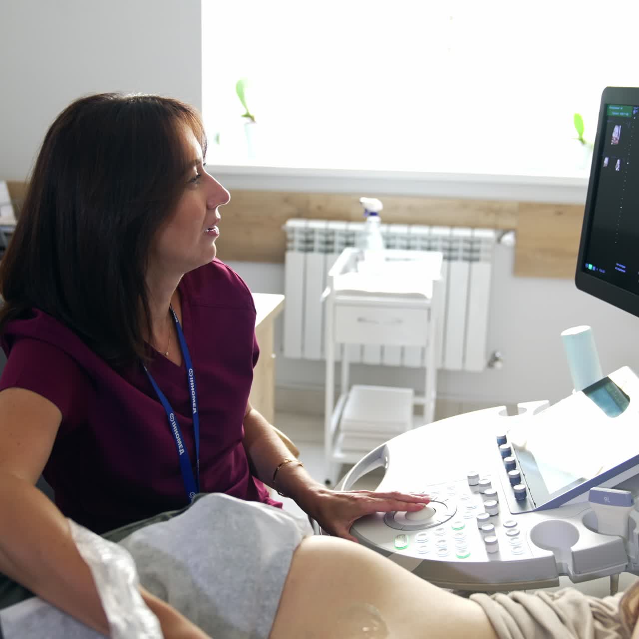 Positive brunette female obstetrician examining the woman at ultrasonic equipment. Doctor is looking at the screen and commenting the process
