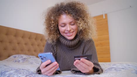 comprador feliz pagando con tarjeta de crédito en línea. mujer blanca rizada usando tarjeta bancaria para el pago en la tienda web en un teléfono inteligente moderno. cliente alegre comprando en línea con teléfono móvil y conexión a internet