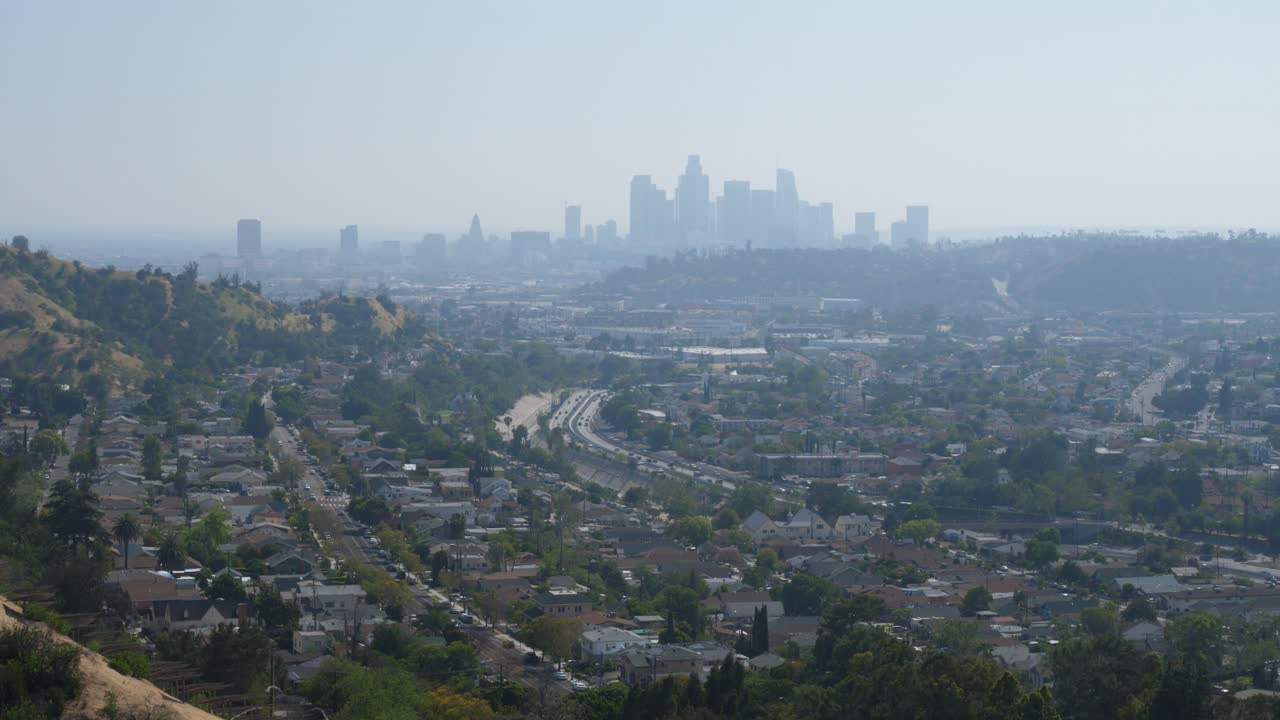 Panoramic view of Los Angeles, with a residential neighborhood in the foreground. The downtown skyline is visible in the distance, with roads and freeways connecting the city