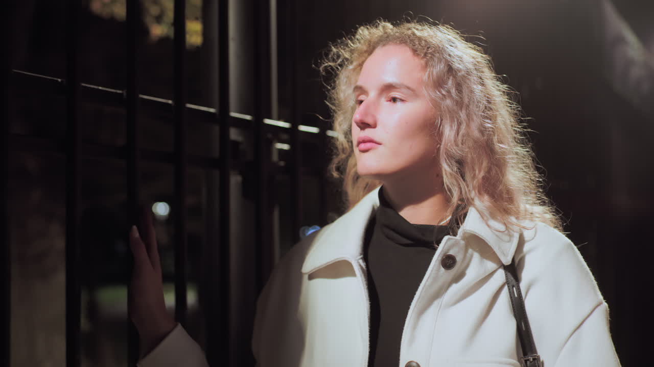 Young lady wearing white coat and black innerwear holding bag strap glides hand on iron fence while walking at night under soft streetlight with calm thoughtful expression and blurred lights behind